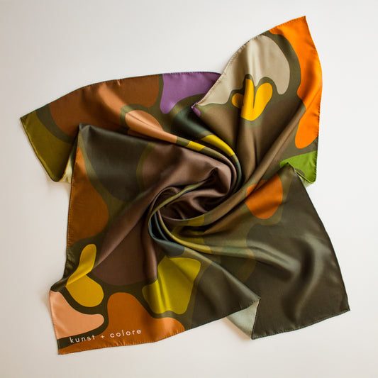 Olaf & Olive Silk Scarf by Kunst & Colore shown arranged artfully on a plain background.  The print features colourful pops of earthy tones on a khaki green background. 