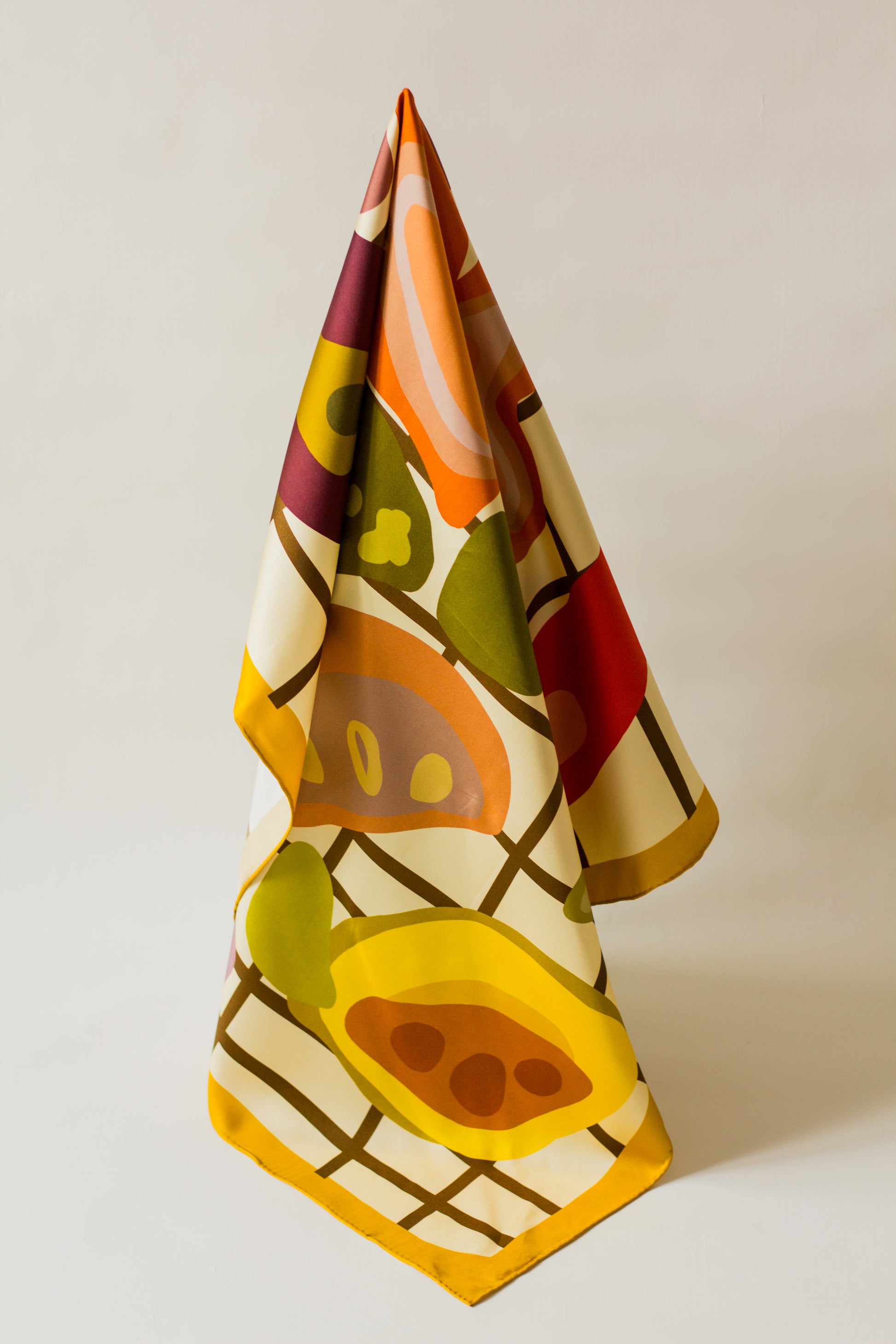 Side Salad by Kunst & Colore silk scarf, hanging artfully on a plain background.  The print features food in earthy tones illustrated on a brown/cream check background, with a mustard yellow border. 