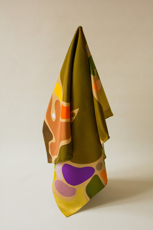 Eat Your Greens Silk Scarf by Kunst & Colore, featuring organic forms coloured in natural tones and bright pops of colour on an oaty beige background.  The scarf hangs artistically on a white background. 