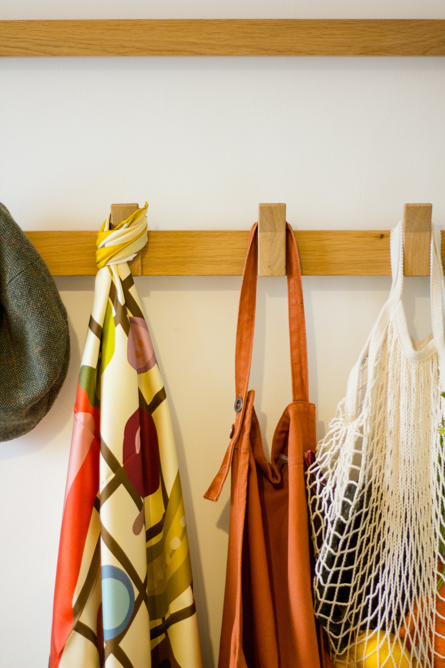 Side Salad by Kunst & Colore silk scarf, shown hanging on coat pegs beside a rust-coloured apron, a string bag of fresh produce, and a tweed flat cap. The print of the scarf features food in earthy tones illustrated on a brown/cream check background, with a mustard yellow border.