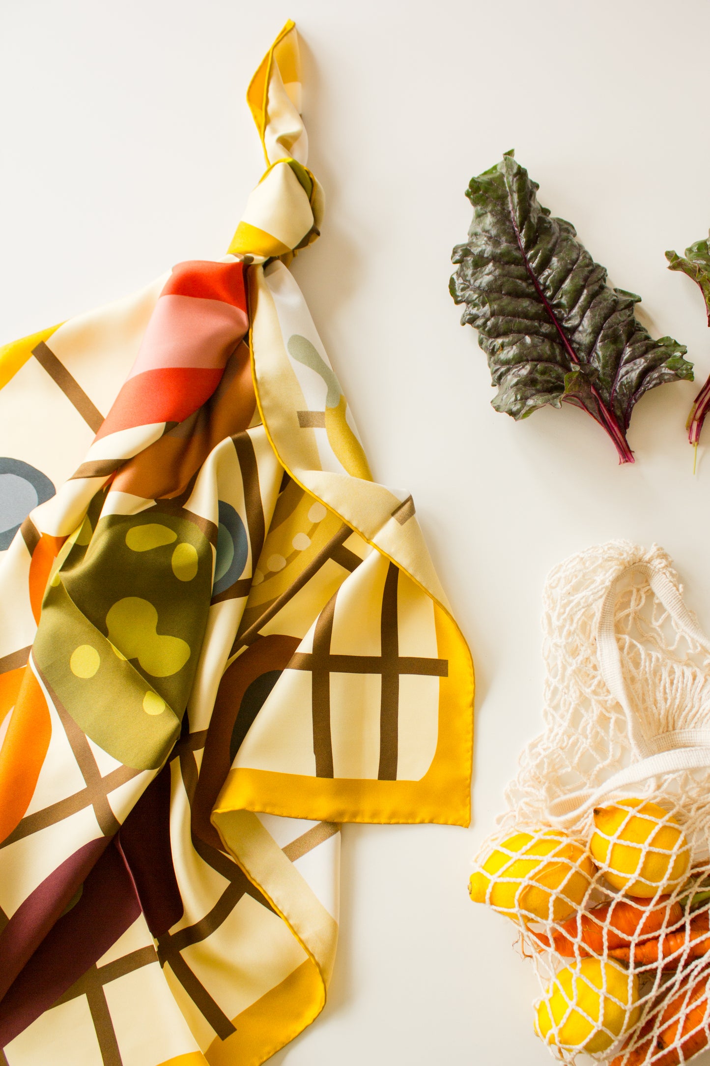 Side Salad by Kunst & Colore silk scarf, tied and arranged artfully next to a string bag of fresh produce.   The print of the scarf features food in earthy tones illustrated on a brown/cream check background, with a mustard yellow border.