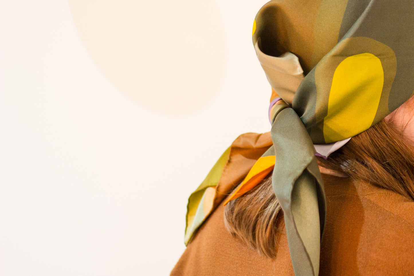 Olaf & Olive Silk Scarf by Kunst & Colore shown tied in a headscarf/bandana style on the models head. The print features colourful pops of earthy tones on a khaki green background.