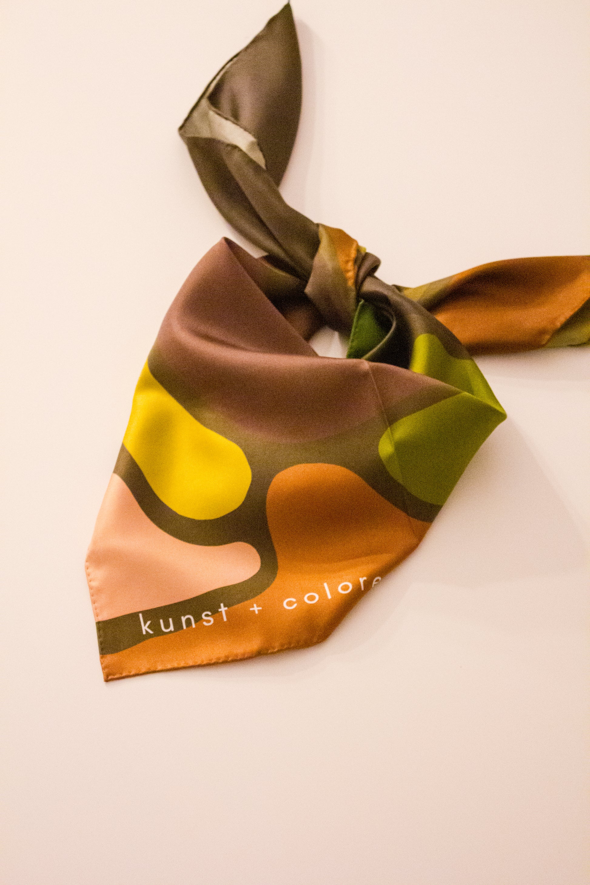 Olaf & Olive Silk Scarf by Kunst & Colore shown tied into a neck scarf shape on a plain background. The print features colourful pops of earthy tones on a khaki green background.