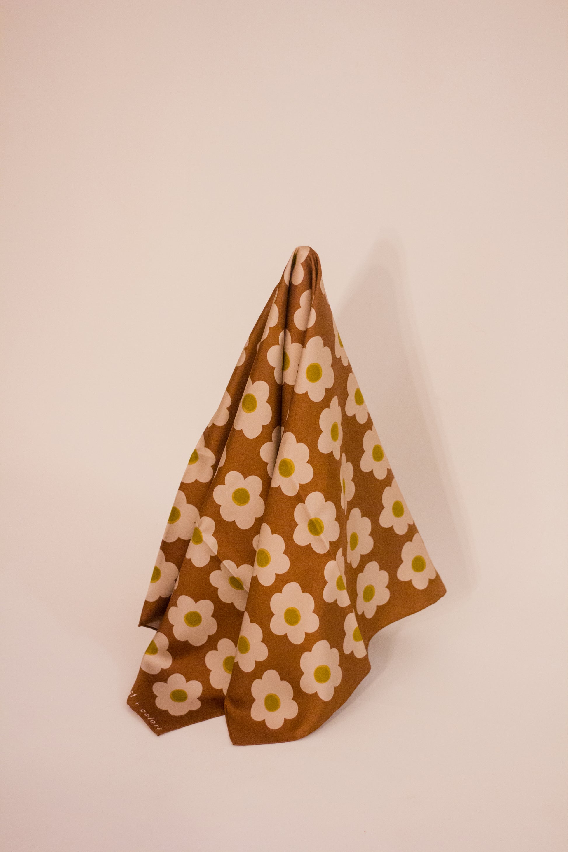The 'Daisy Brown' scarf by Kunst & Colore hanging artfully. The print features small cream-coloured daisies arranged in a uniform square, on a brown background. 