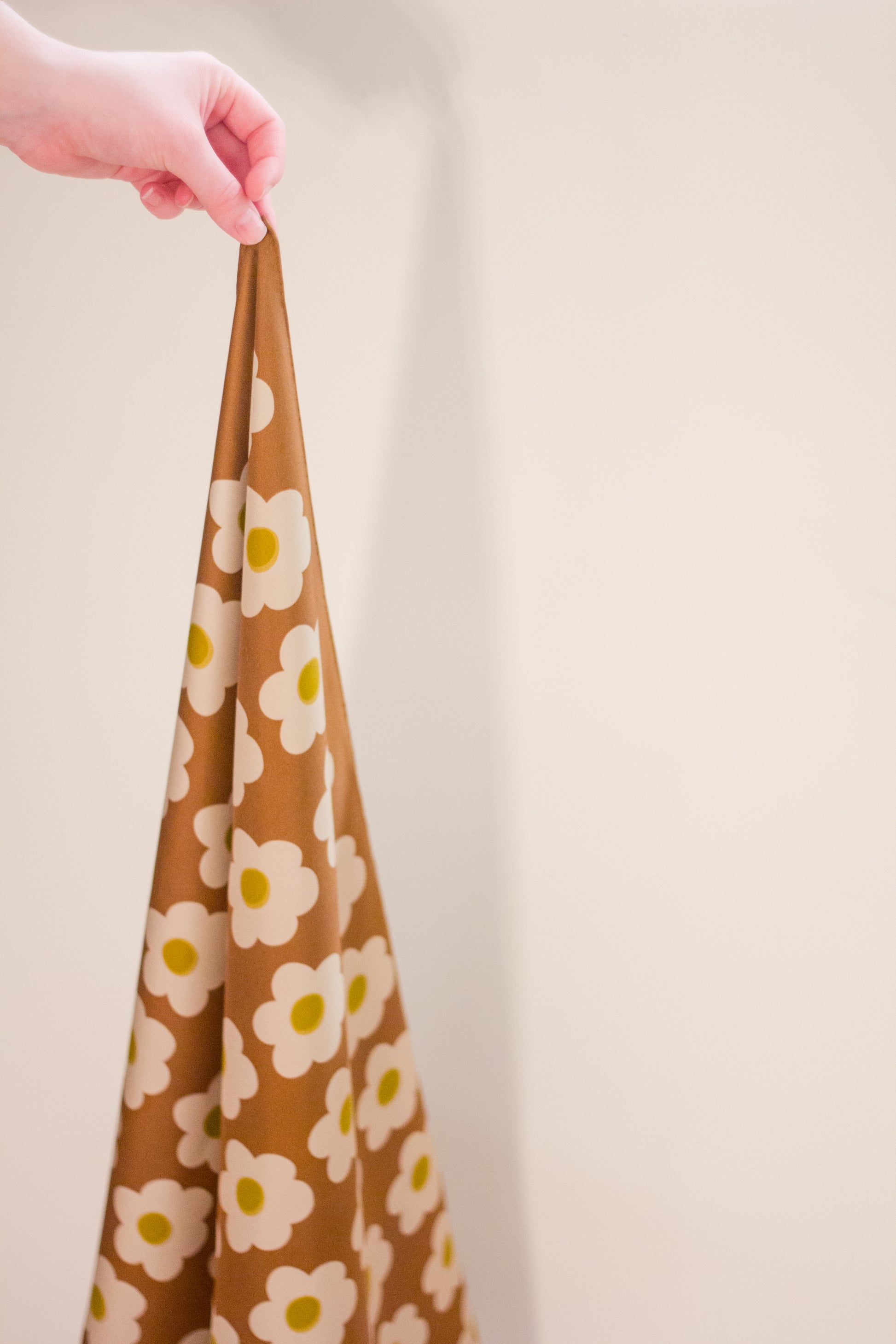 A hand holding the 'Daisy Brown' scarf by Kunst & Colore as it hangs beneath artfully. The print features small cream-coloured daisies arranged in a uniform square, on a brown background. 